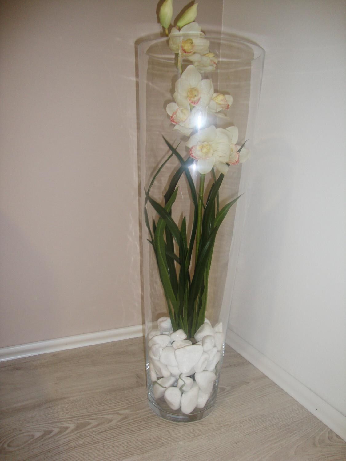 A clear glass vase with white flowers and green leaves on a wooden floor.