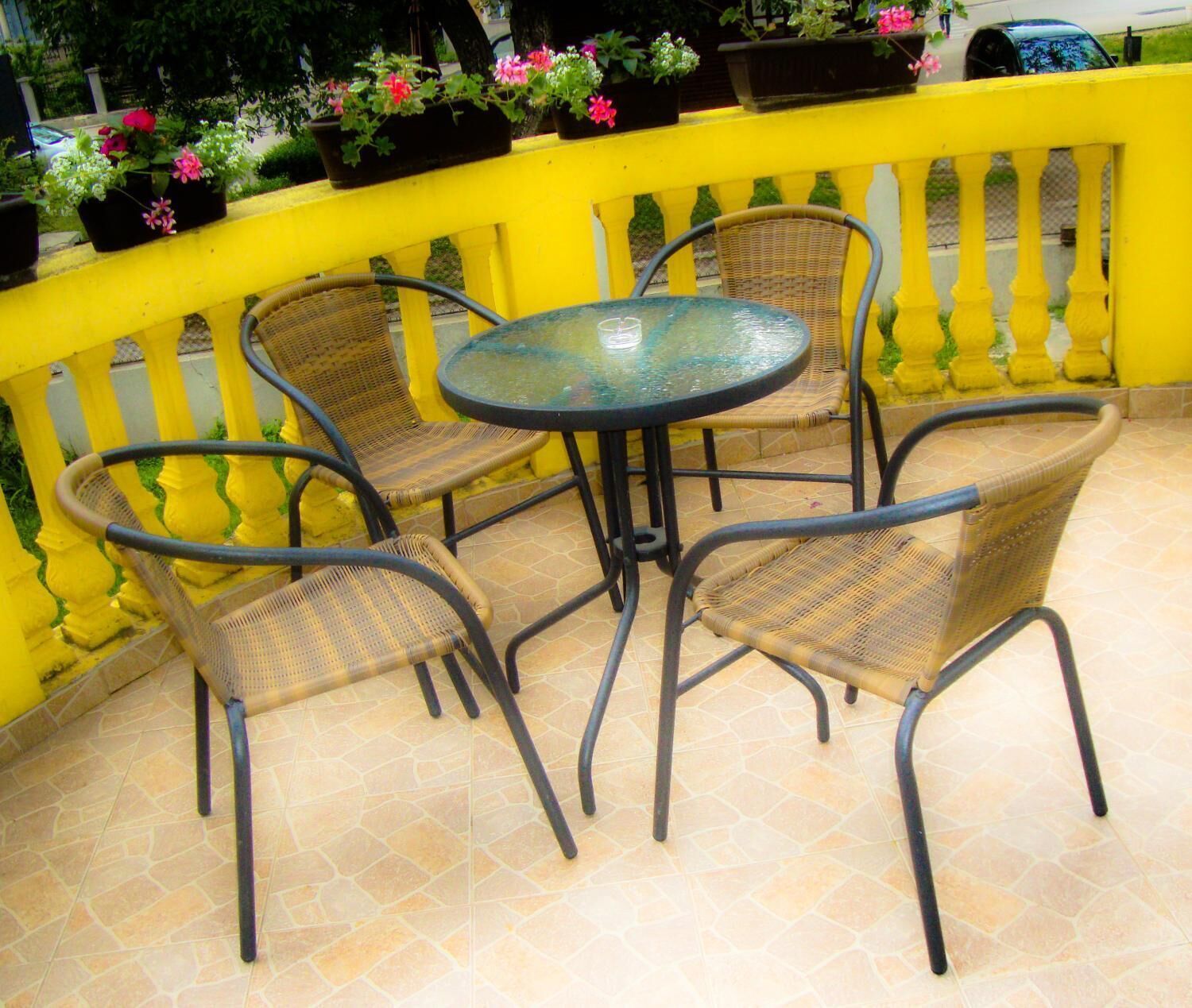 Een terras met een ronde tafel en vier rieten stoelen, omgeven door een gele reling.