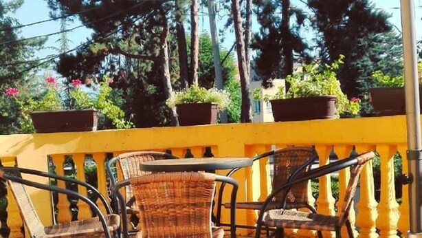 Een terras met rieten stoelen en een tafel, een gele balustrade en potplanten.