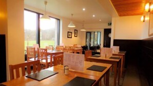 A dining area with wooden tables and chairs, large windows, and pendant lighting. - An Cala Cafe and Bunkhouse - Hostel (Lochinver)