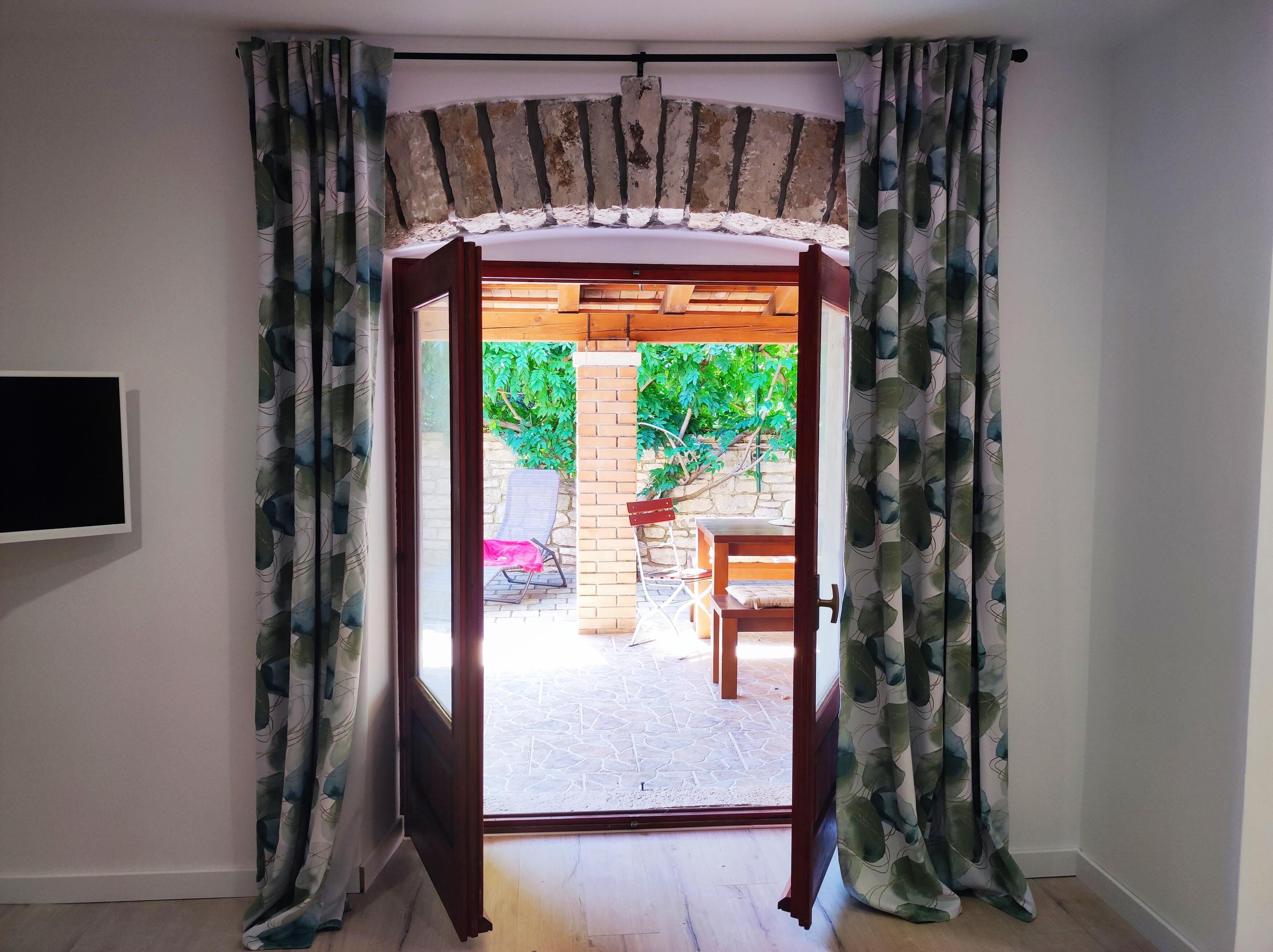 Una puerta que da a un patio con una mesa y sillas de madera, un muro de piedra y follaje.