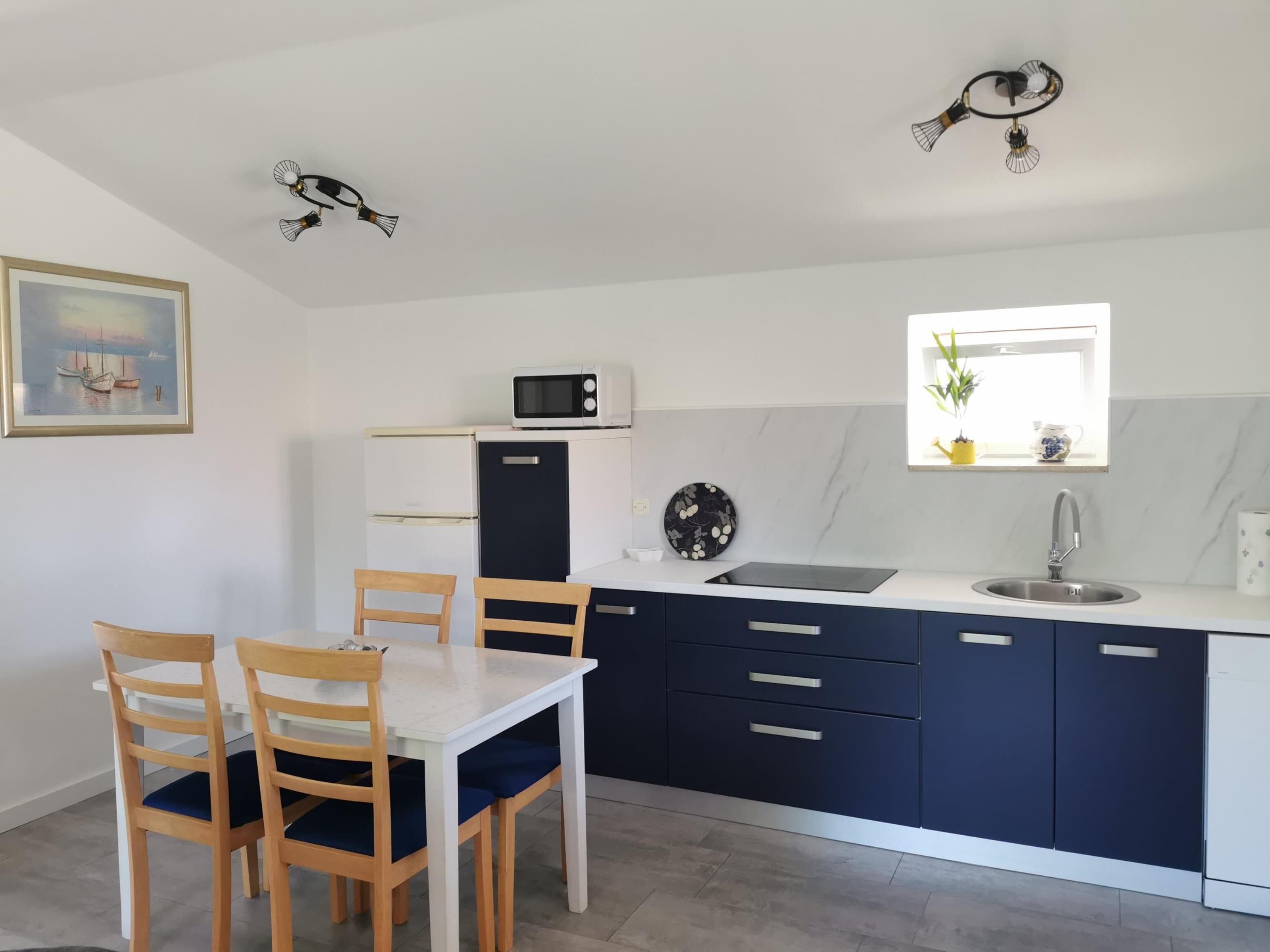 A compact kitchen with a white countertop, dark blue cabinets, a microwave, a sink, and a window with a plant.