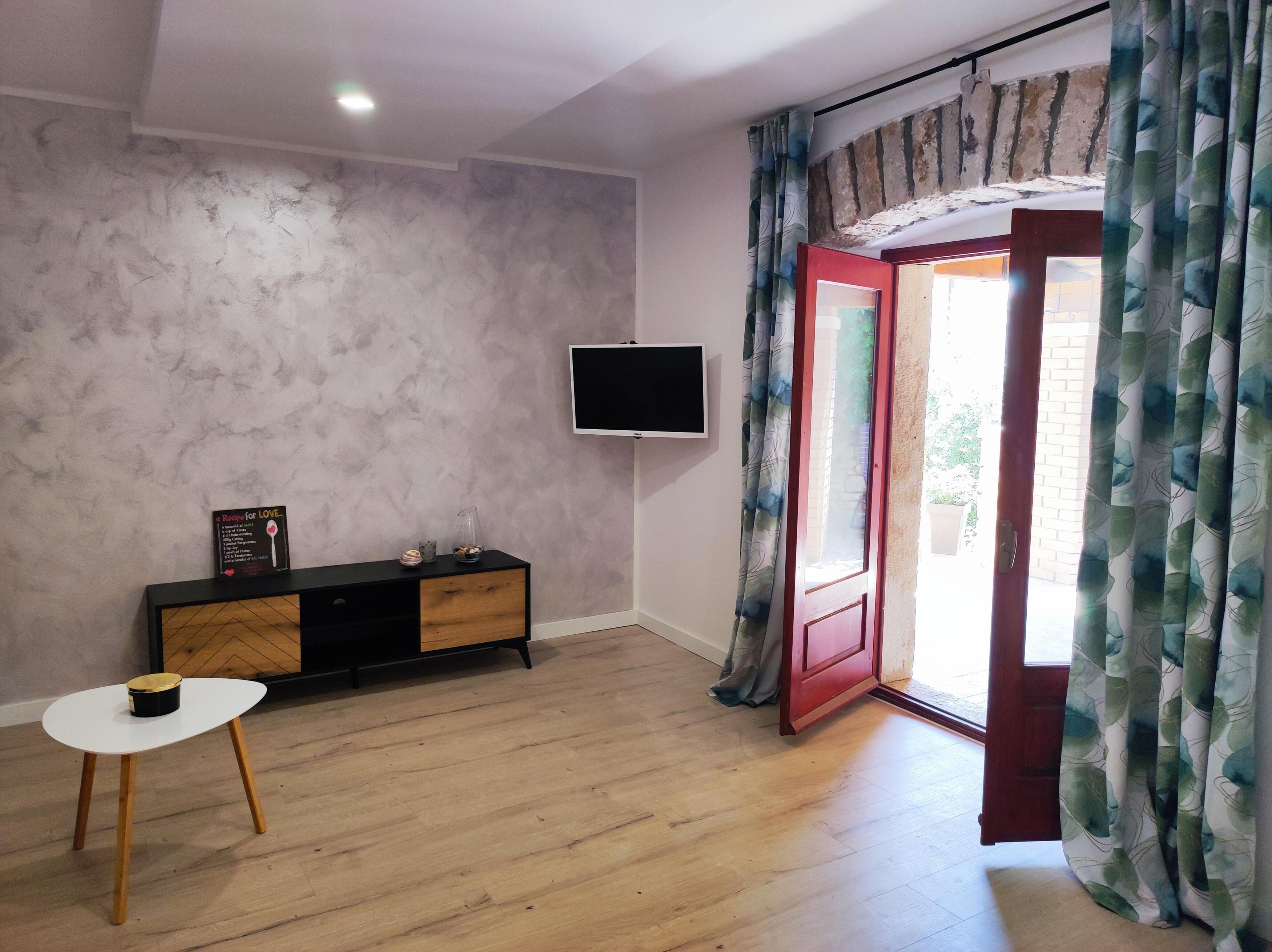 A room with a wooden floor, a black TV on the wall, a small white table, and a red door with patterned curtains.