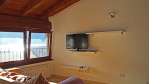 Una habitación con techo de madera, una ventana con persianas, una cama con manta, un televisor montado y un termostato.