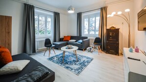 Un salón moderno con un sofá gris, un sillón negro y una mesa de centro redonda. Hay dos ventanas con cortinas, un radiador y un mueble con un espejo.