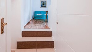 Un pasillo que conduce a un sofá azul frente a una pared blanca, con un cuadro enmarcado encima del sofá.