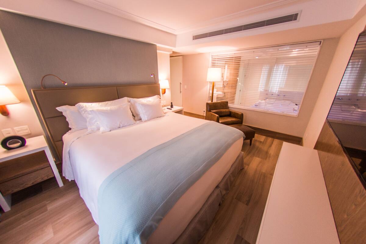 Una habitación de hotel con una cama grande, mesitas de noche, una silla y una ventana con persianas.