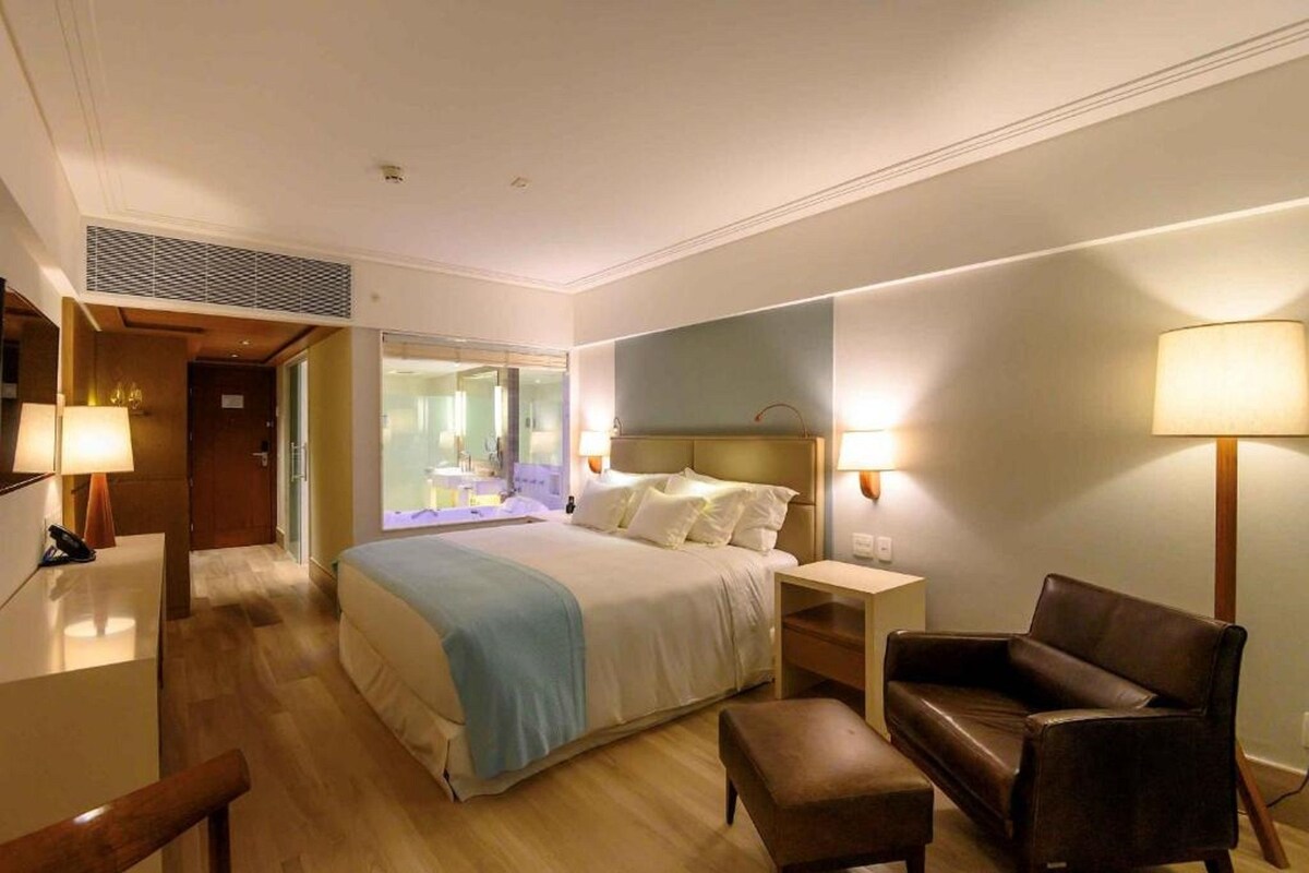 Una habitación de hotel con cama, mesitas de noche, una silla, un escritorio y una lámpara.