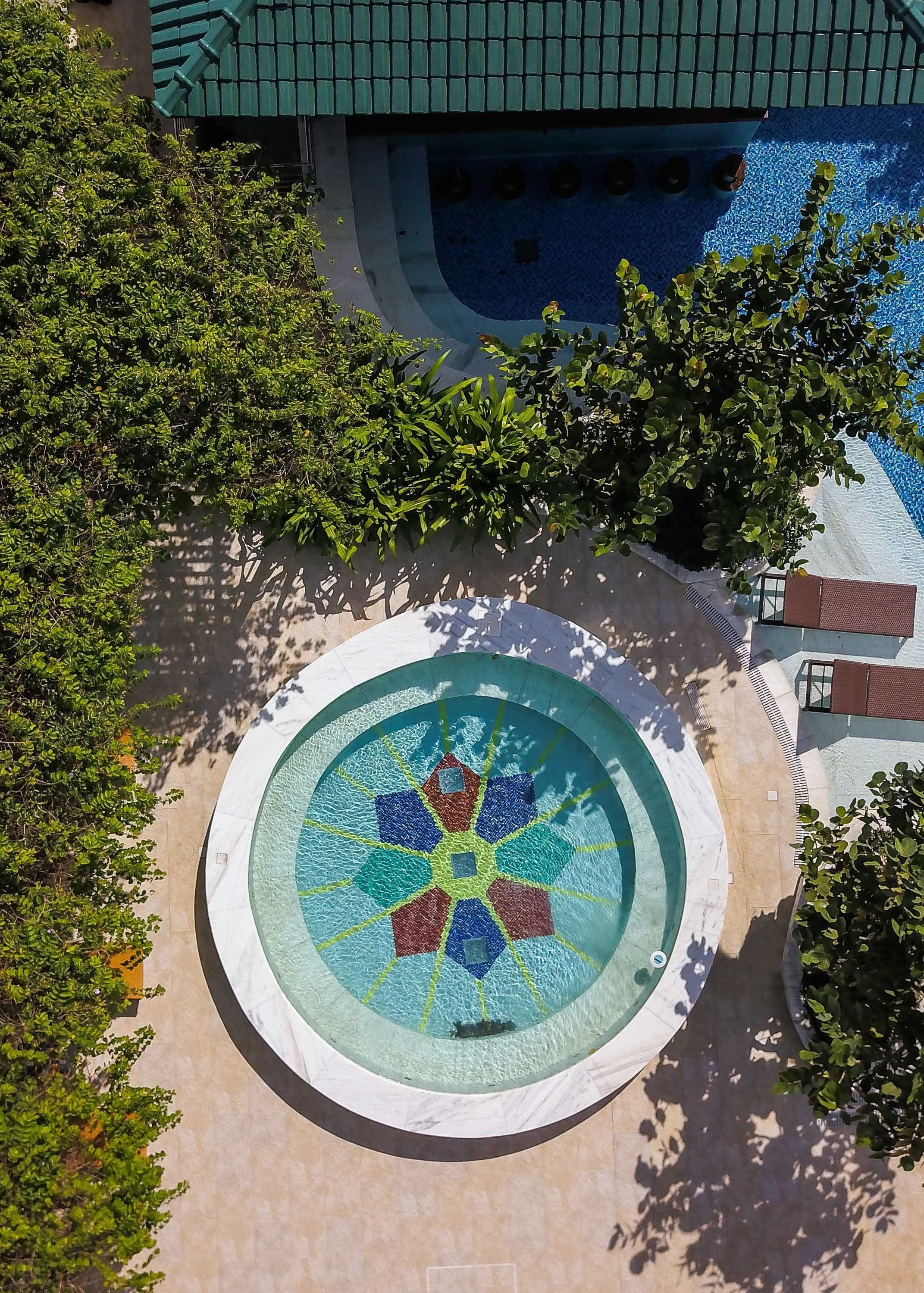 Uma piscina circular com design em mosaico, rodeada por árvores e um edifício com telhado verde.