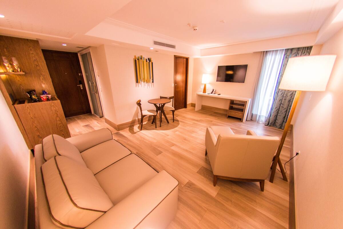Una habitación de hotel moderna con un sofá, una silla, una mesa de comedor con sillas, un televisor montado en la pared y una lámpara.