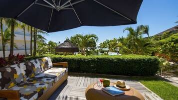 A wooden deck with a sofa and a round wicker ottoman, a large umbrella, and a view of a pool and palm trees.