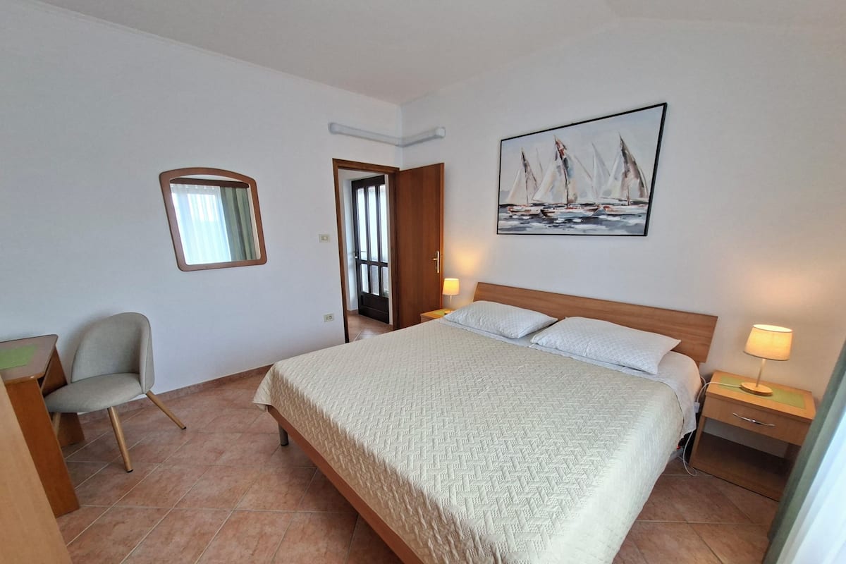 Une chambre d’hôtel avec un lit, une chaise, un bureau, une table de chevet, une lampe, un tableau et une fenêtre.