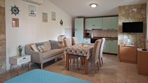 A living room with a sofa, a dining table with chairs, a kitchen area with cabinets, a television mounted on a stone wall, and an air conditioning unit on the wall. - Apartments Marlina (Rovinj)