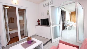 Ett kompakt bostadsutrymme med en skjutdörr i glas som leder ut till en balkong, ett litet pentry, en TV och en sittgrupp med en rosa soffa.