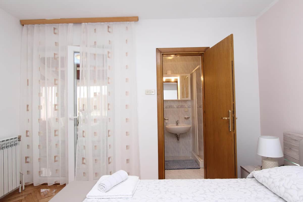 A bedroom with a bed, a bedside table with a lamp, a wooden door leading to a bathroom, and sheer curtains.