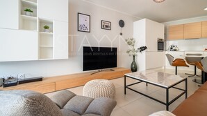 A modern living room with a flat-screen TV, a coffee table, and a sofa. There is a kitchen area with wooden cabinets and a dining table with chairs. - Boutique 2 bedroom apartment VRBO (Nice)