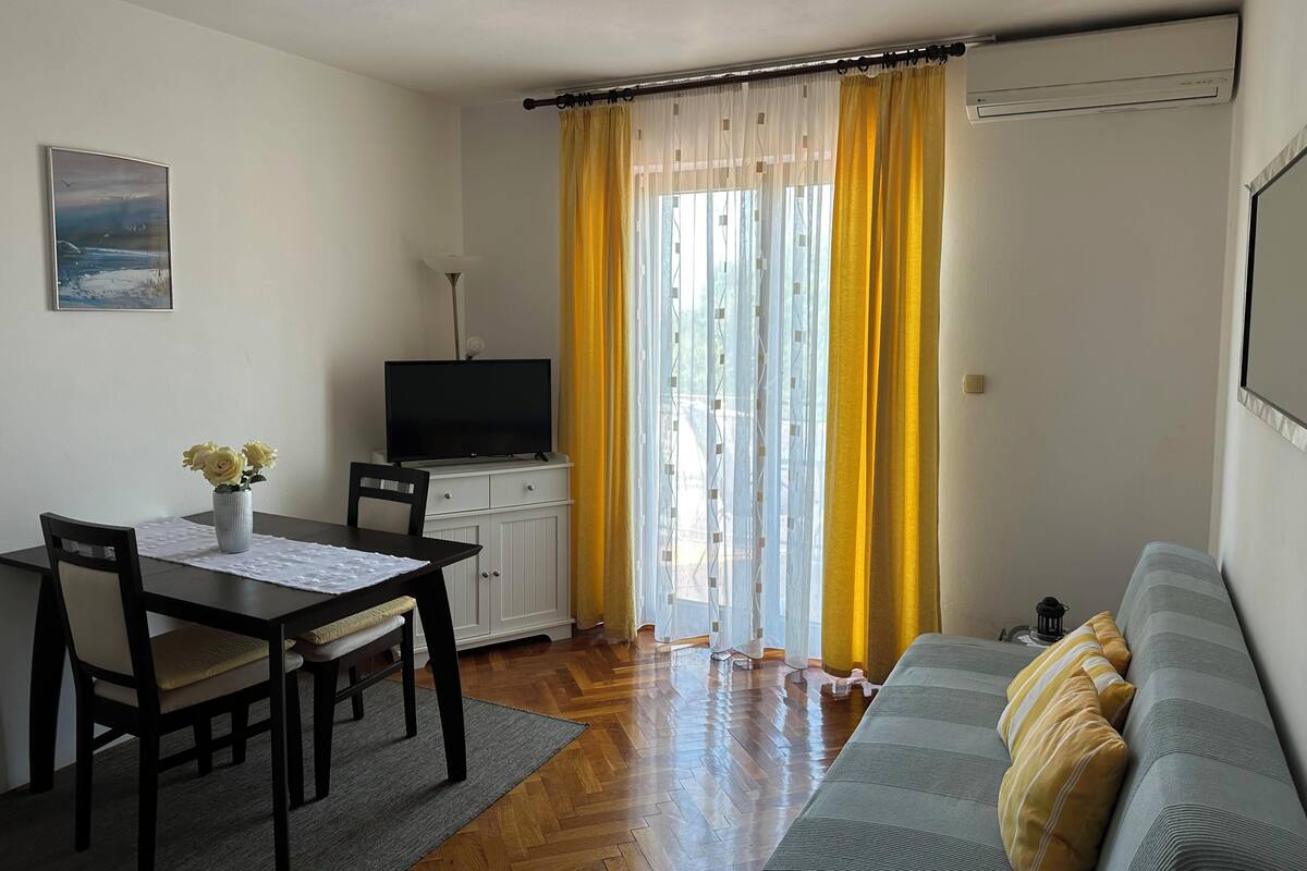 Un salon avec un canapé, un téléviseur posé sur un meuble, une table à manger avec des chaises et une fenêtre ornée de rideaux jaunes.