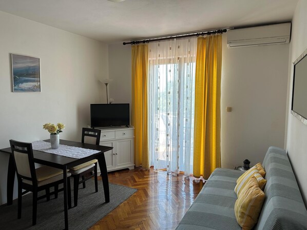 Una sala de estar con un sofá, un televisor sobre un mueble bajo, una mesa de comedor con sillas y una ventana con cortinas amarillas.