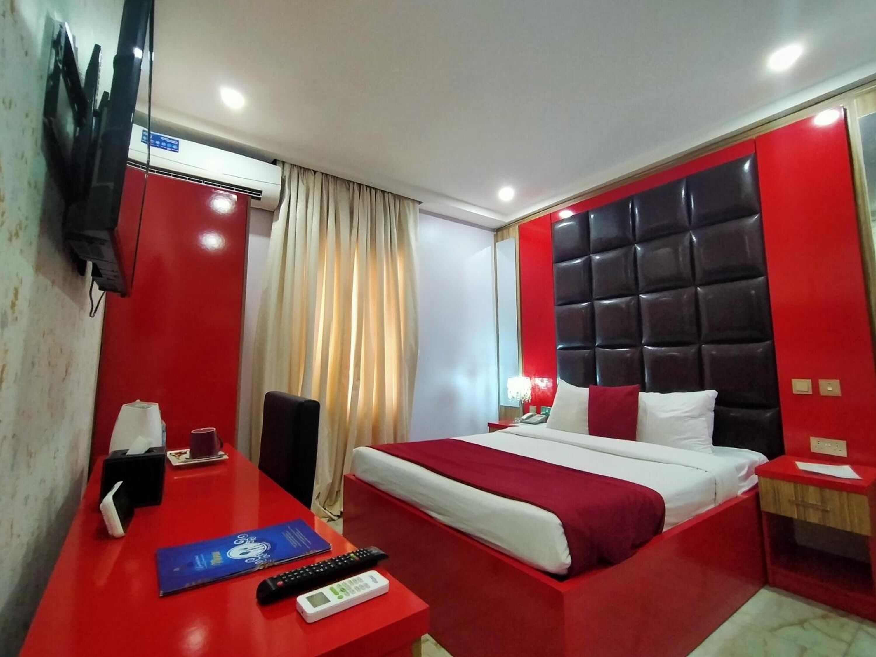 A hotel room with a red and white color scheme, a large bed with a black headboard, a desk with a telephone and remote controls, and a mounted TV on the wall.
