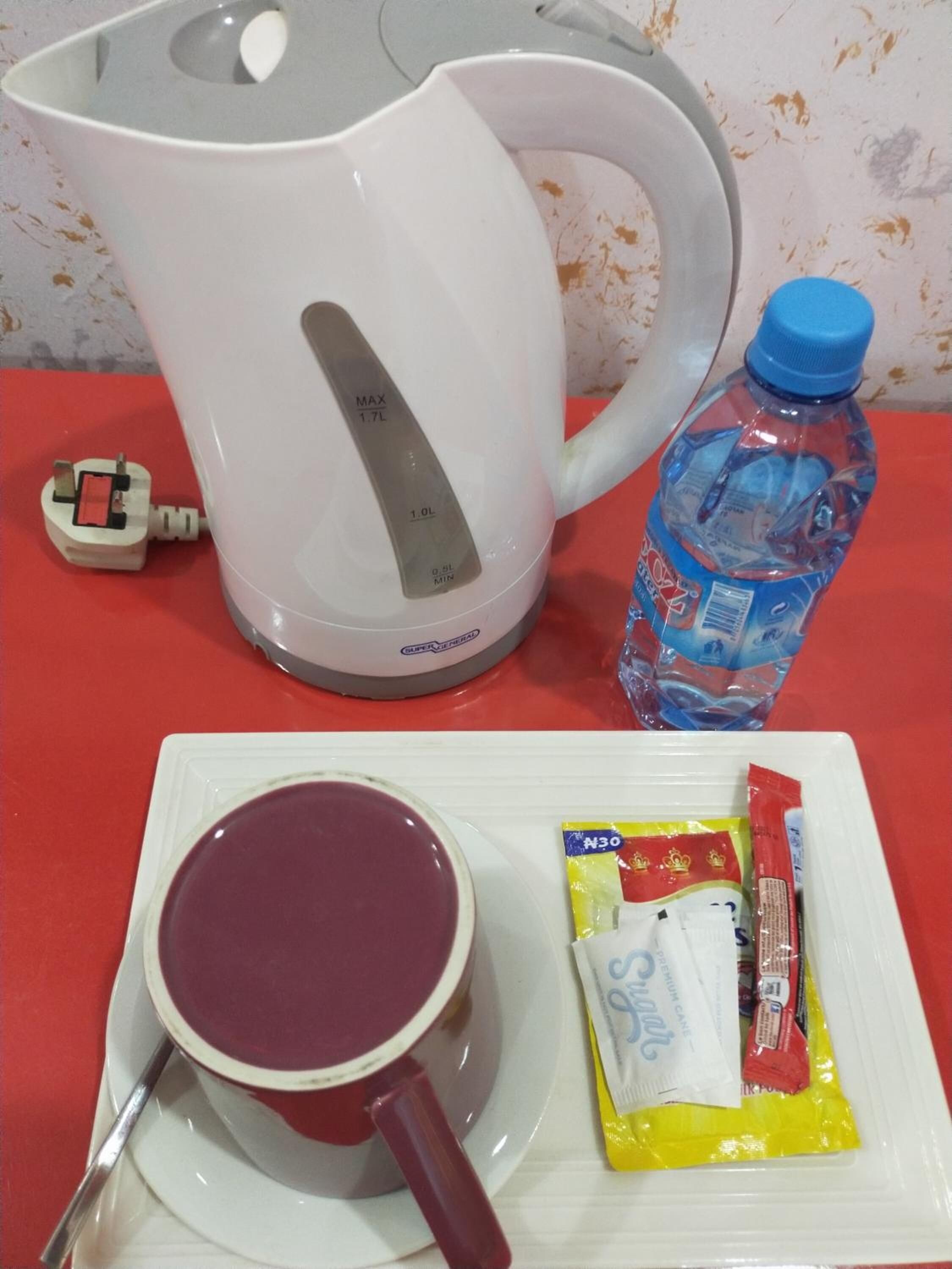 A white electric kettle, a water bottle, a mug with a spoon, and packets of snacks on a red table.