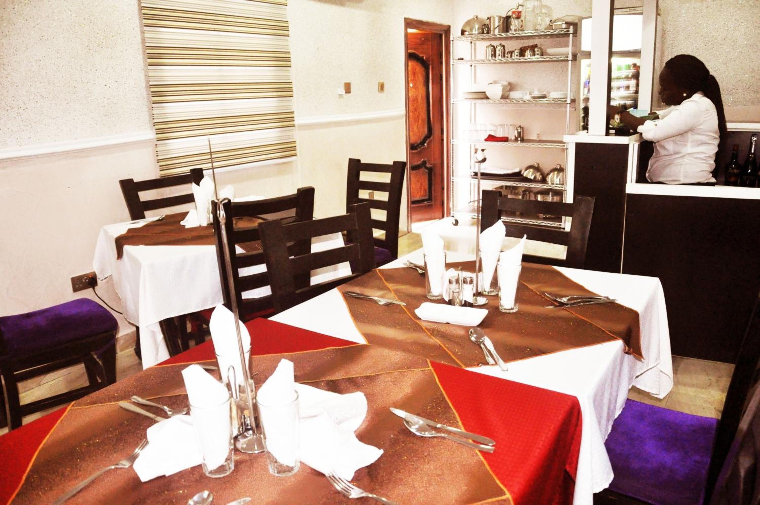 Un comedor con mesas dispuestas para cuatro, cada una con mantel blanco, posavasos rojos y cubertería. Se observa una barra en el fondo, atendida por una persona.