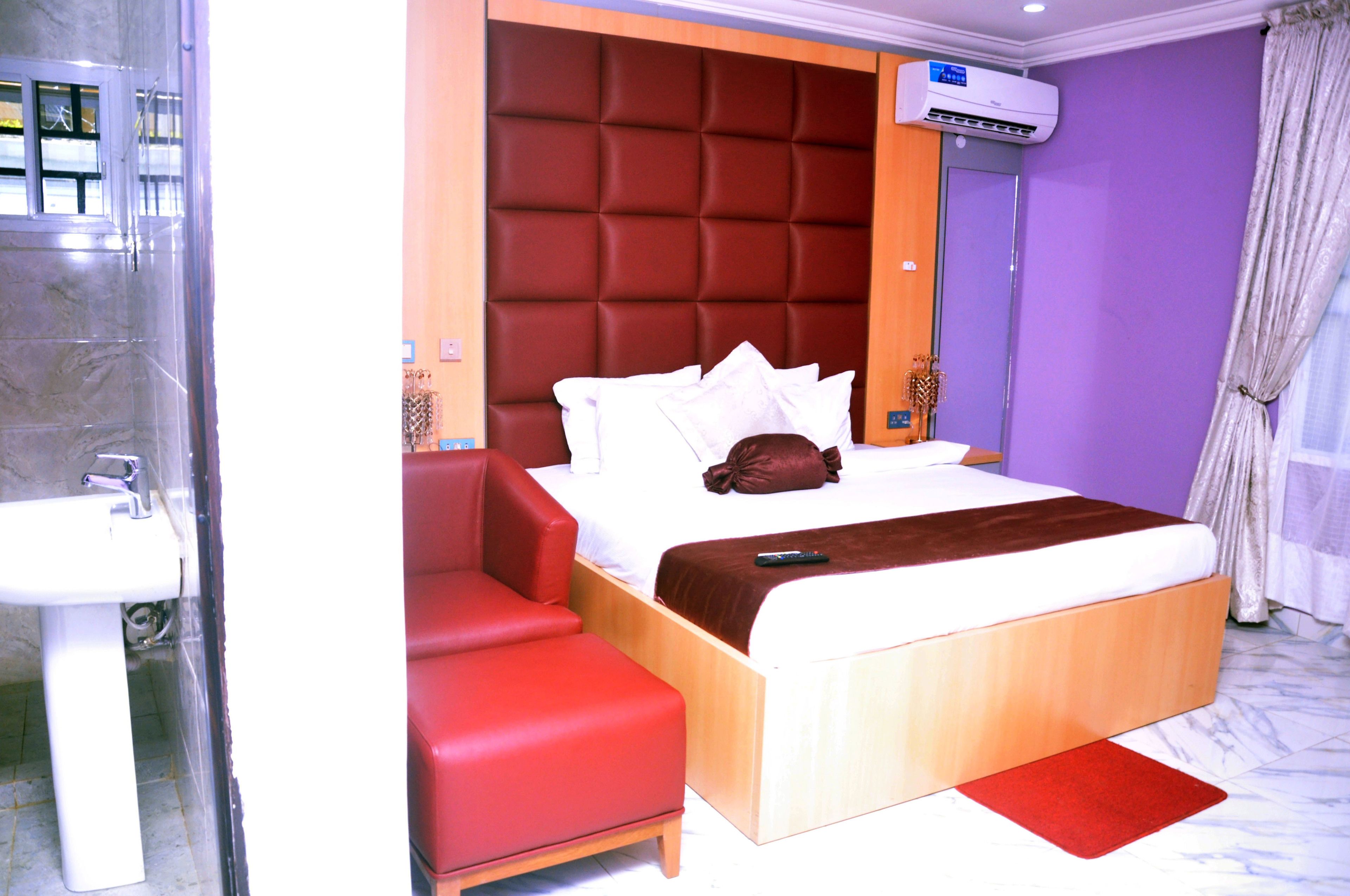 Une chambre d’hôtel avec un lit, une chaise rouge, une table de chevet en bois, un lavabo blanc et un appareil de climatisation.