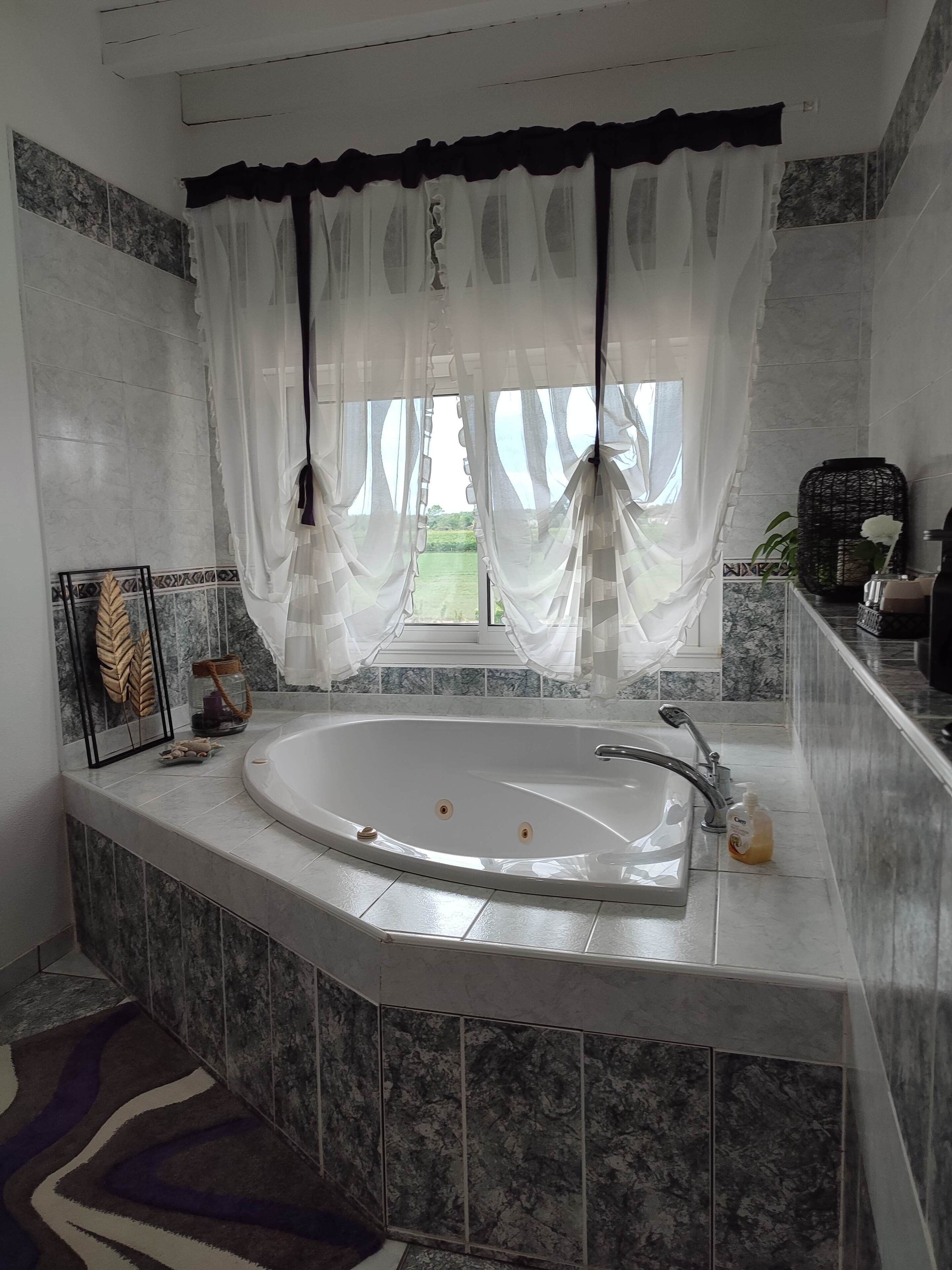 A bathroom with a freestanding bathtub, a window with sheer curtains, and a tiled backsplash.