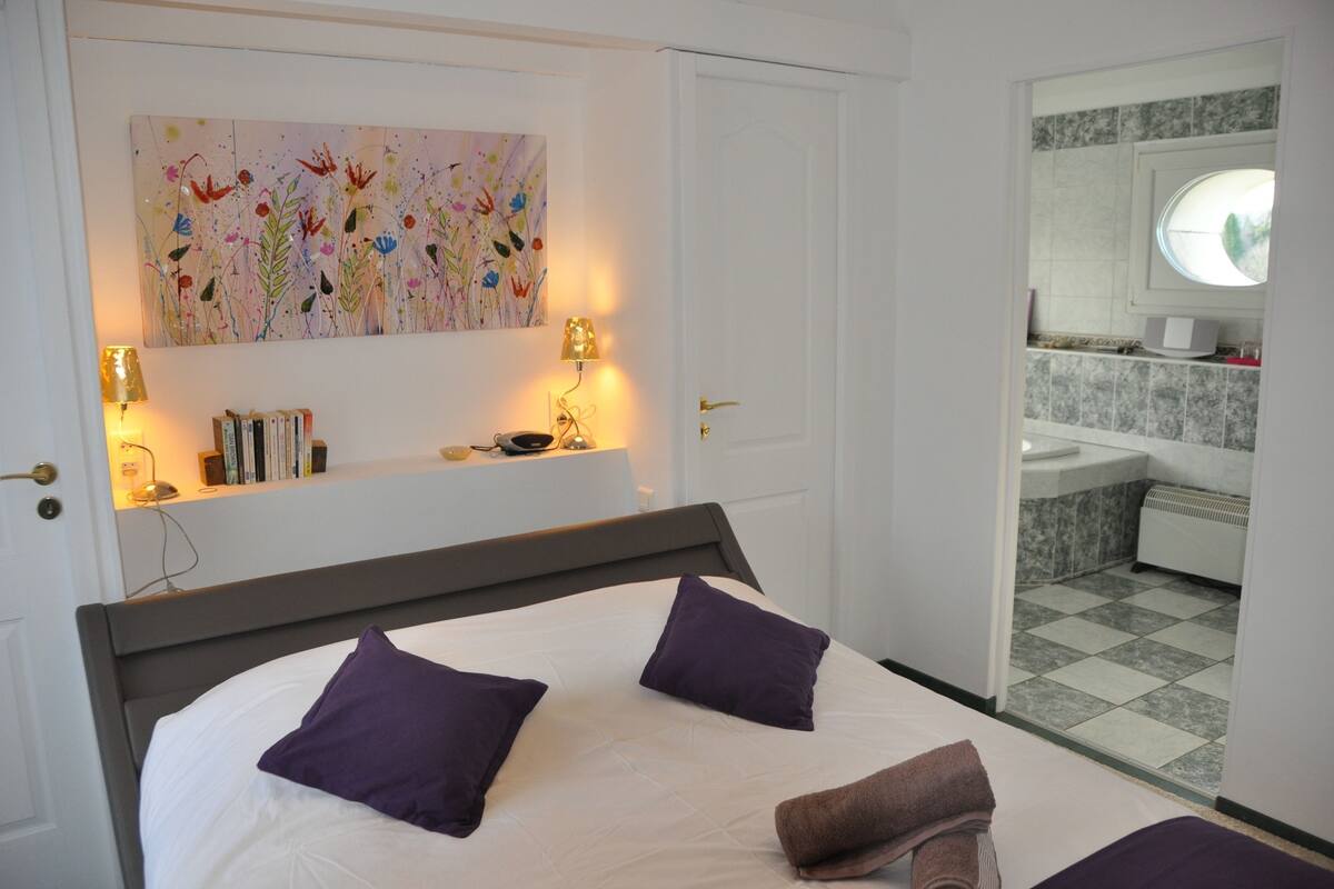 Une chambre à coucher bien rangée, avec un lit, des oreillers, des lampes de chevet, une bibliothèque et un tableau accroché au mur.