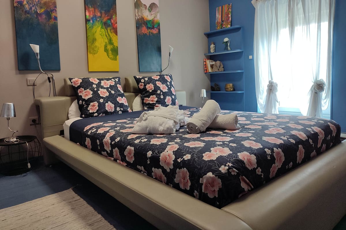 Une chambre à coucher avec un grand lit, une literie à motifs floraux, une table de chevet, une lampe et des œuvres d’art accrochées aux murs.