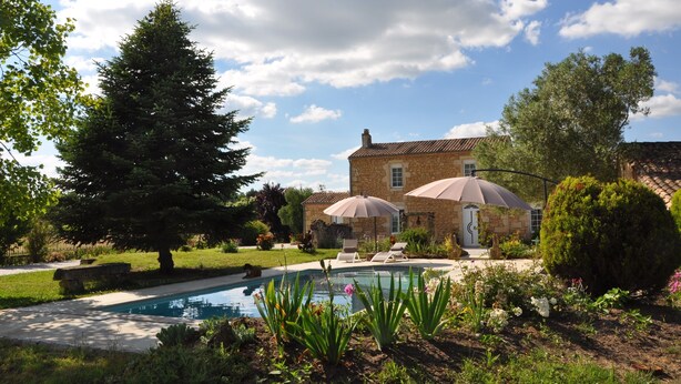 A stone house with a swimming pool, surrounded by lush greenery and a clear sky.
