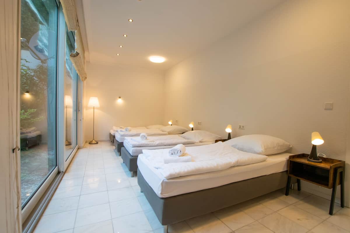 A hotel room with four beds aligned along a glass wall, each with a bedside lamp.