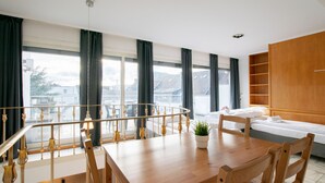 Une chambre d’hôtel moderne avec une table à manger en bois, des chaises, un canapé et une grande fenêtre avec des rideaux.