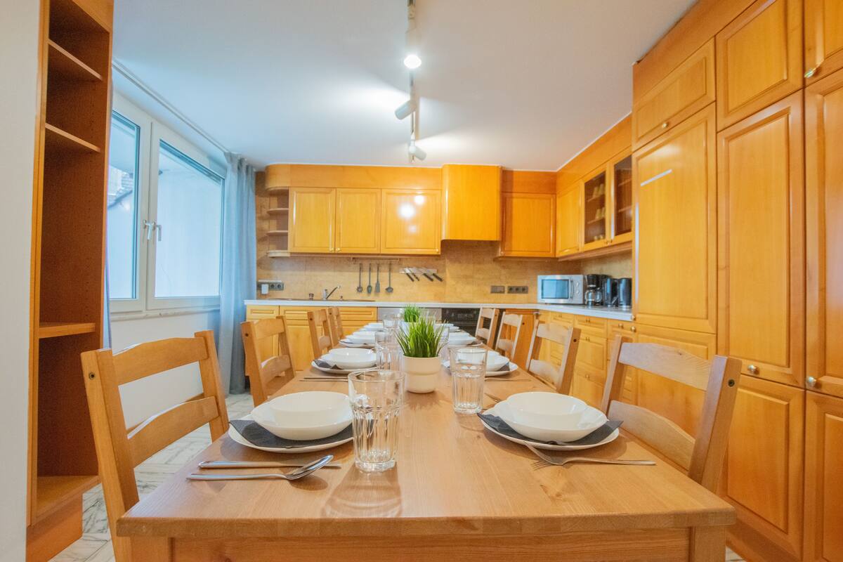 A dining area with a wooden table set for six, surrounded by wooden chairs. There is a window on the left side, and kitchen cabinets are visible in the background.