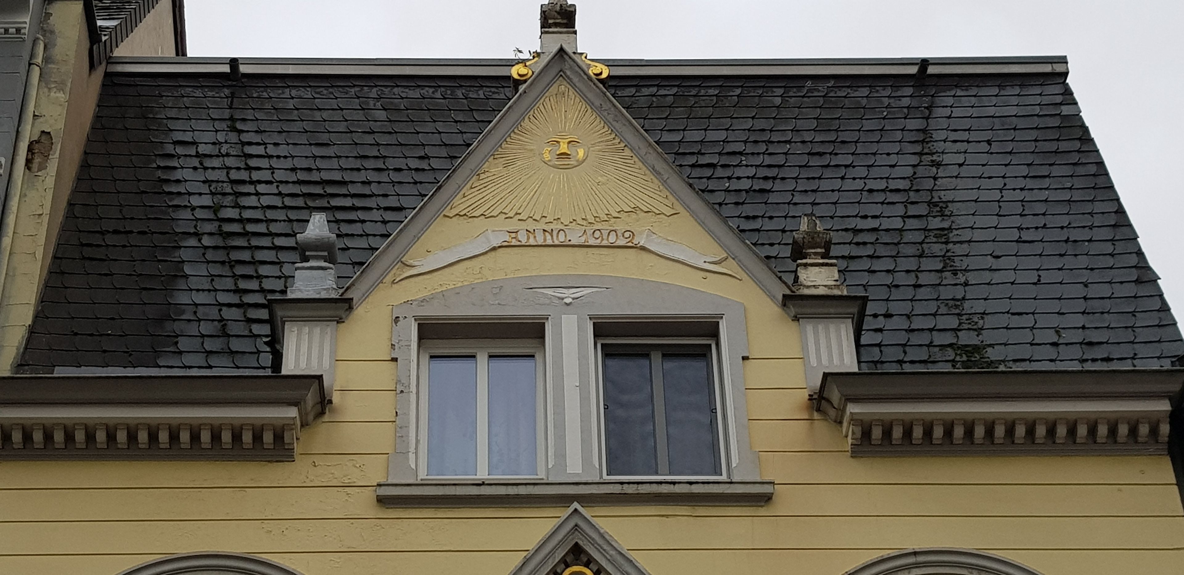 A building with a yellow facade, a black tiled roof, and a triangular gable with a decorative emblem.