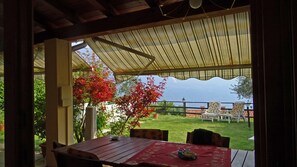 Eine überdachte Terrasse mit Tisch und Stühlen, Blick auf das Meer und ein Garten mit roten Blättern.