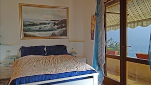 Ein Schlafzimmer mit einem Bett, einem Meeresblick-Gemälde, einem Nachttisch, einer Lampe und Meerblick durch eine Glastür.