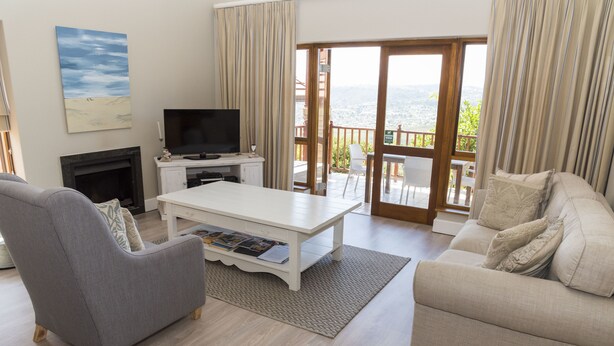 A living room with a fireplace, a flat-screen TV, a white coffee table, a grey armchair, and a beige sofa. - Villa 12 (Knysna)