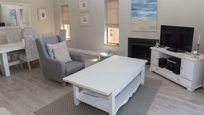 A living room with a grey sofa, white coffee table, and a TV on a white cabinet. There are framed pictures on the wall and a fireplace. - Villa 12 (Knysna)