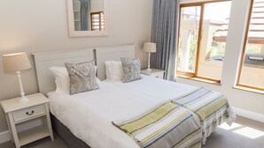 A neatly made bed with a striped blanket, white bedding, and decorative pillows. There is a bedside table with a lamp and a small drawer. A window with curtains is visible, and a mirror hangs on the wall above the bed. - Villa 12 (Knysna)