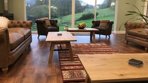 A spacious living room with a large window offering a view of a green landscape. There are two leather sofas, a wooden coffee table, and a rug. - Godshill Park Barn (Ventnor)