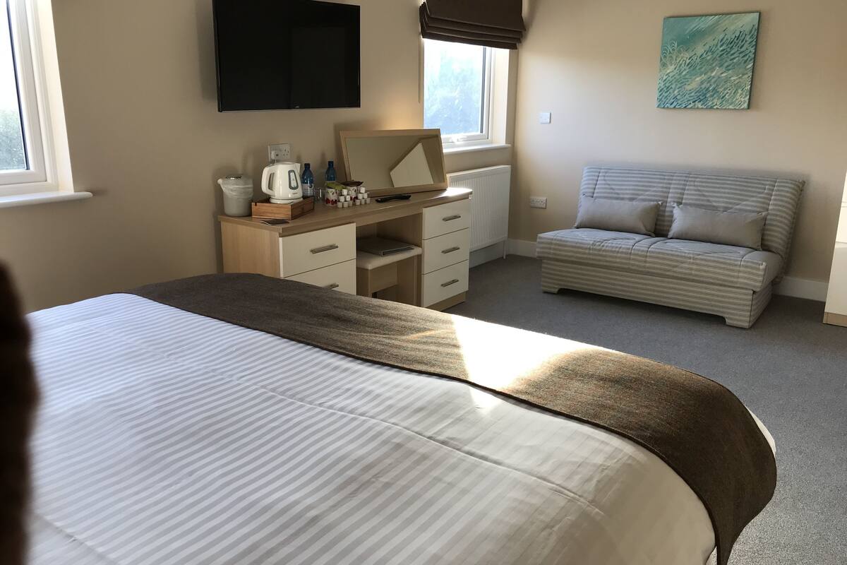 Hotellihuone, jossa on sänky, työpöytä tietokoneella, sohva ja taulu seinällä.