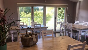 A dining area with a wooden table and white chairs, a wicker basket with a tag, and a view of greenery through large windows. - Godshill Park Barn (Ventnor)