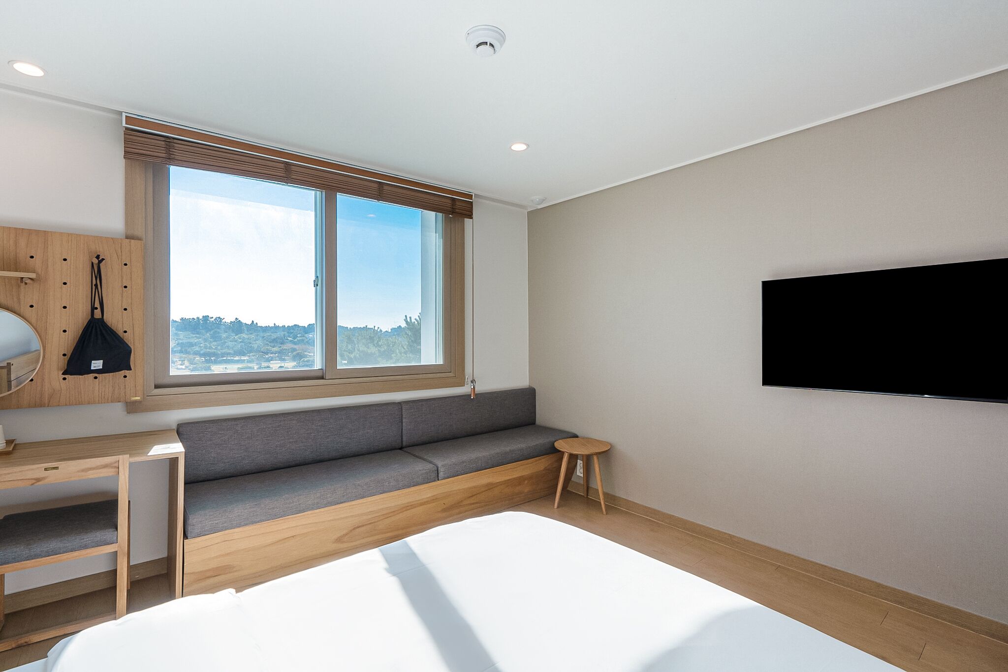A modern hotel room with a large bed, a built-in desk, a grey sofa, a small round table, a TV mounted on the wall, and a window with a view of the city.