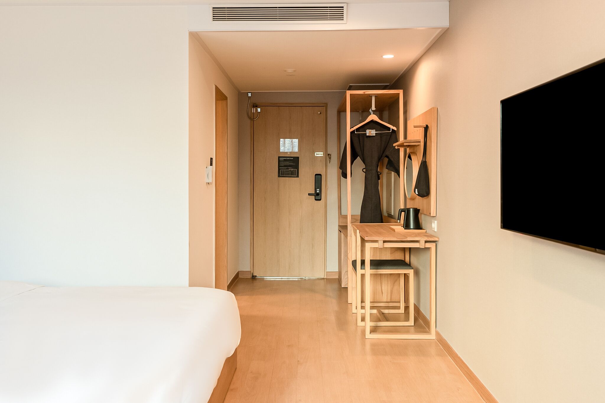 Una camera d'albergo con un letto, una scrivania in legno con una sedia, un appendiabiti e una televisione a parete.