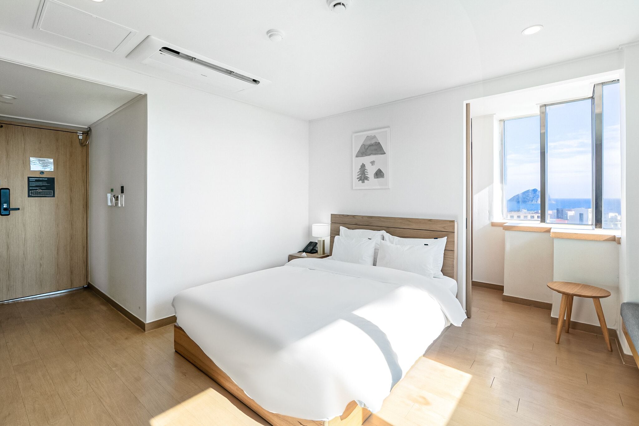 A modern hotel room with a large bed, a wooden floor, a small table, a nightstand, and a window with a view.