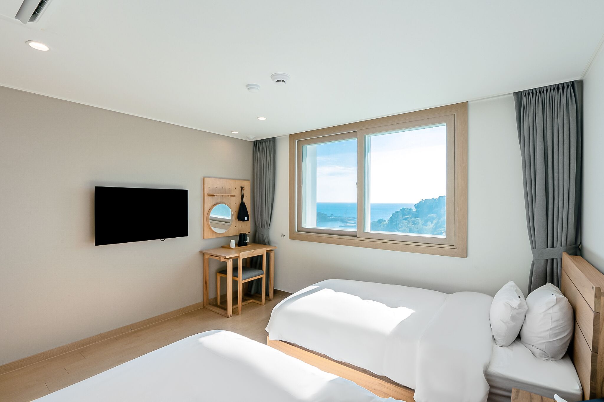 A hotel room with two beds, a TV, a desk, and a window with a view of the sea.
