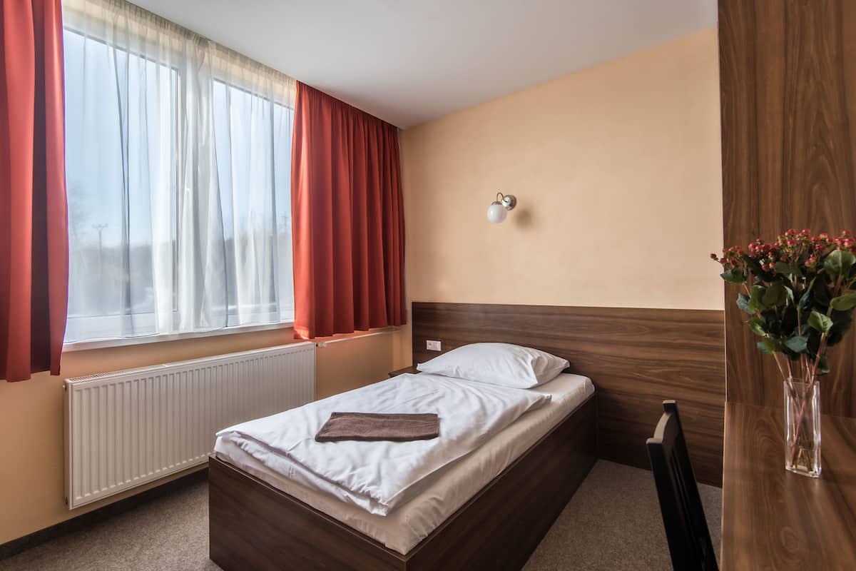 A neatly made bed with white linens and a brown throw, a bedside table with a vase of flowers, and a window with red curtains.