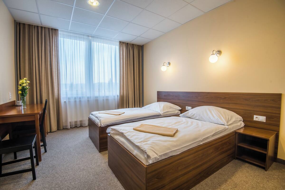 A hotel room with two single beds, a wooden desk with a chair, a small nightstand, and a window with curtains.