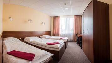A hotel room with three single beds, a wooden wardrobe, a desk, and a large window with curtains.
