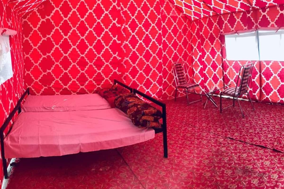 Quarto com parede estampada em vermelho e branco, uma cama de solteiro com colchão rosa, uma janela e duas cadeiras.